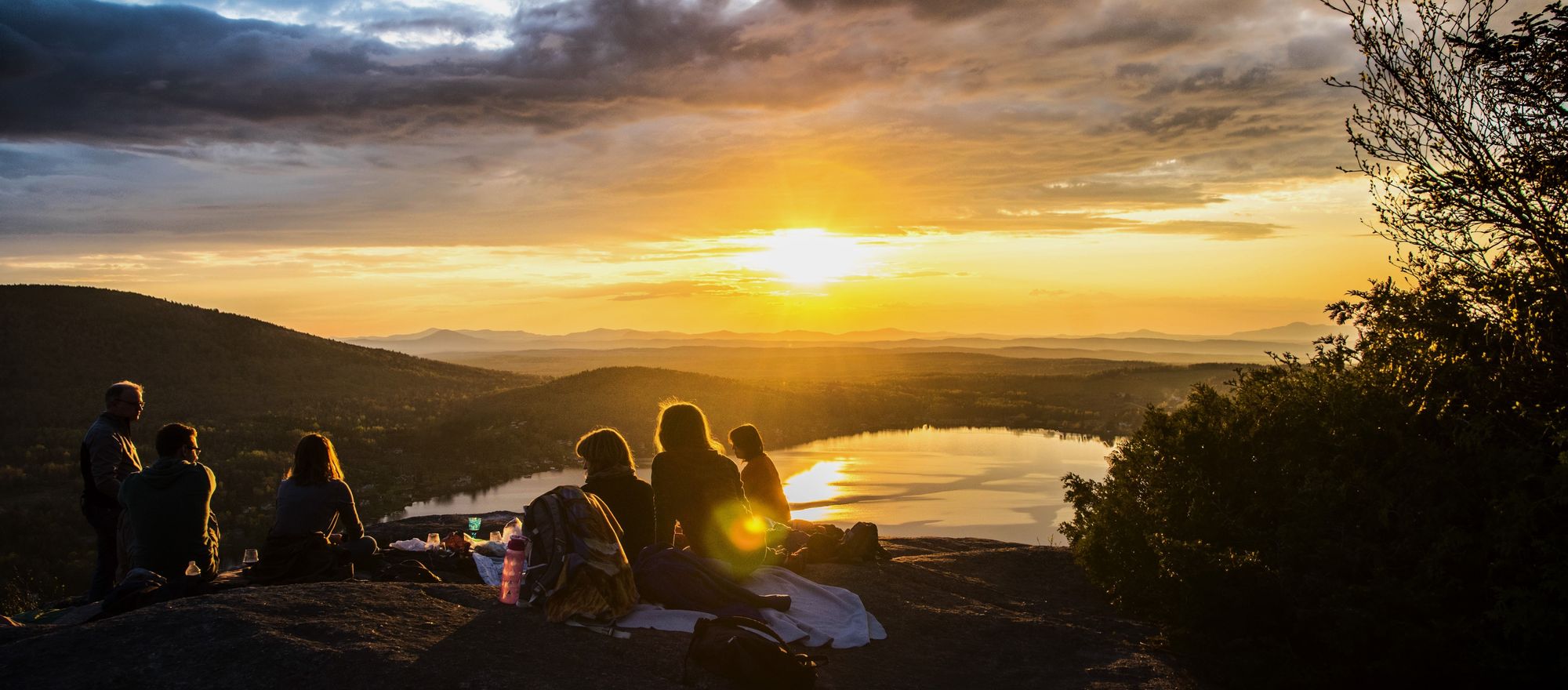A group of people gathered on a hill watching the sun hiding under clouds behind a mountains and lake landscape
